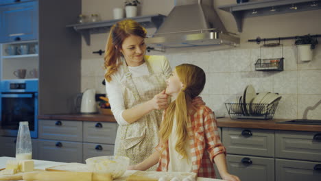 Portrait-shot-of-the-young-mother-and-her-cute-daughter-standing-closely,-looking-at-each-other-and-thanto-the-camera-in-the-kitchen-at-the-table-while-baking.-Indoors
